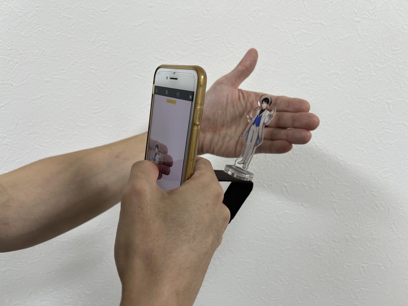 アクスタの裏に手のひらを添えて、iPhoneでピントを合わせているところ。画面に「AE/AFロック」と表示されている。